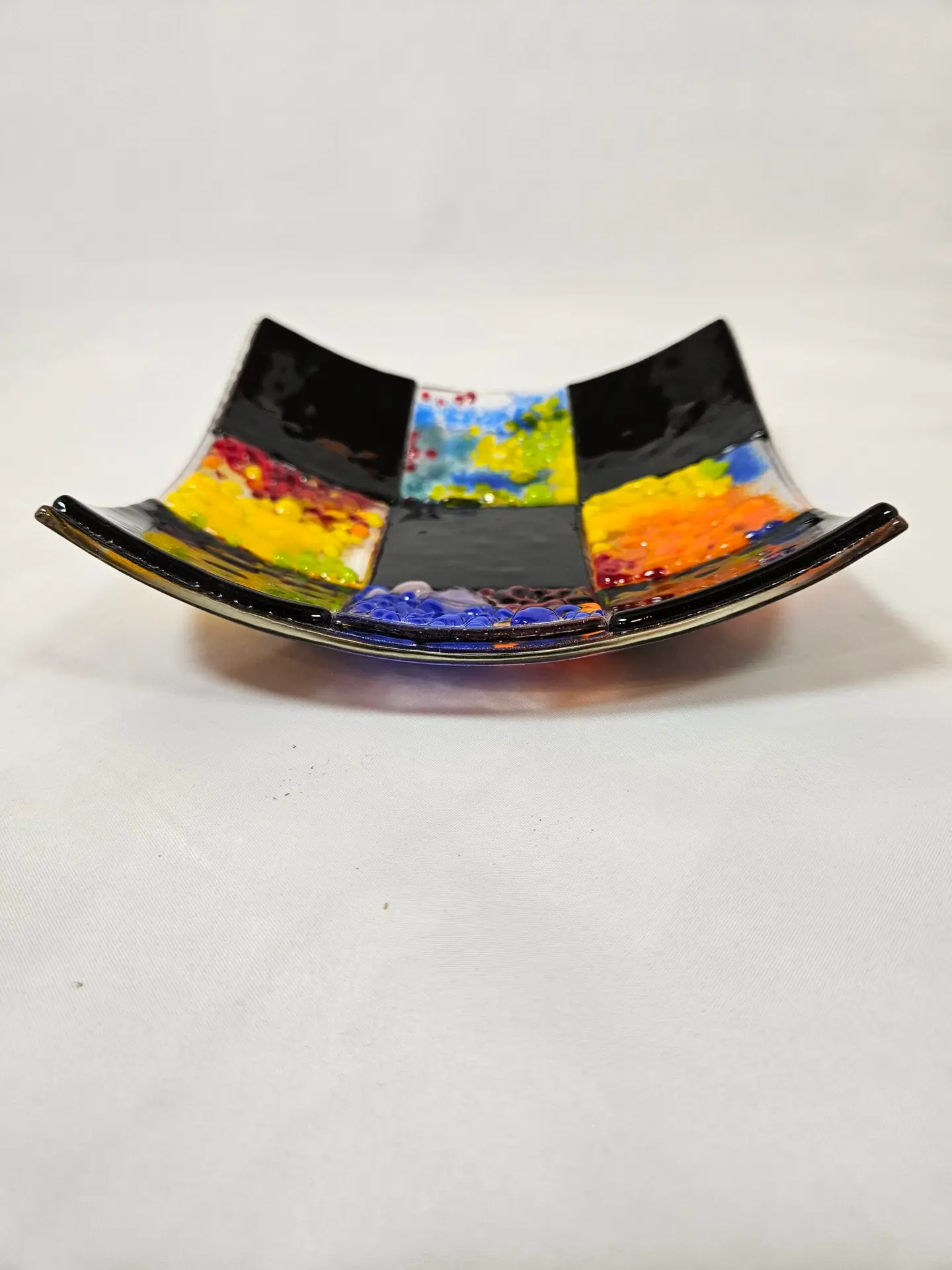 The Patchwork in Black 2 is a square glass dish with a unique curved design, showcasing a vibrant geometric pattern of black, red, yellow, and blue squares.
