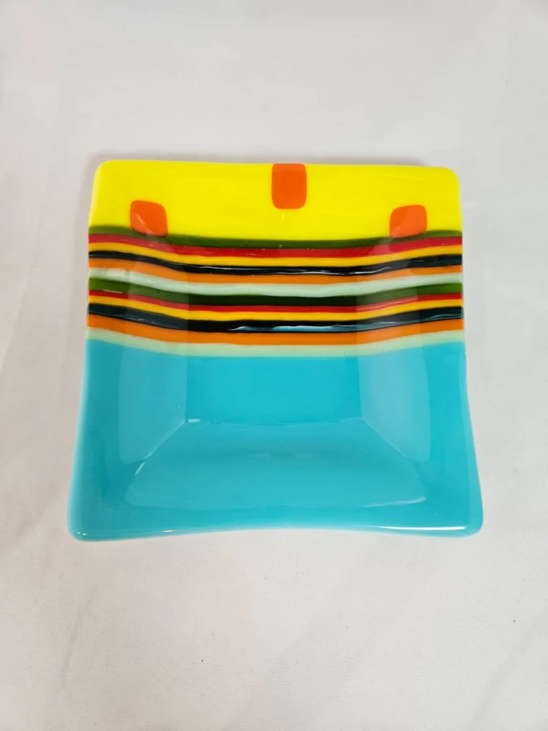 The Striped Radiance plate is a decorative square with a colorful design: the top section features yellow with three orange squares, followed by black, green, yellow, and red stripes. The bottom half is solid light blue. It rests on a plain white surface.