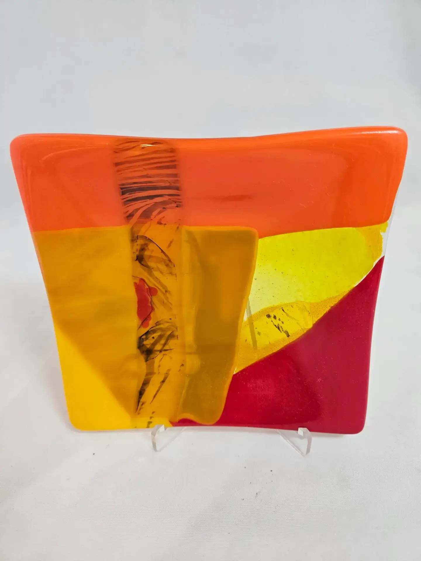 The Radiant Horizon is a vibrant glass art piece with an abstract design, showcasing bold sections of orange, red, yellow, and touches of black and white. It has a glossy finish and is displayed on a clear stand against a plain white background.