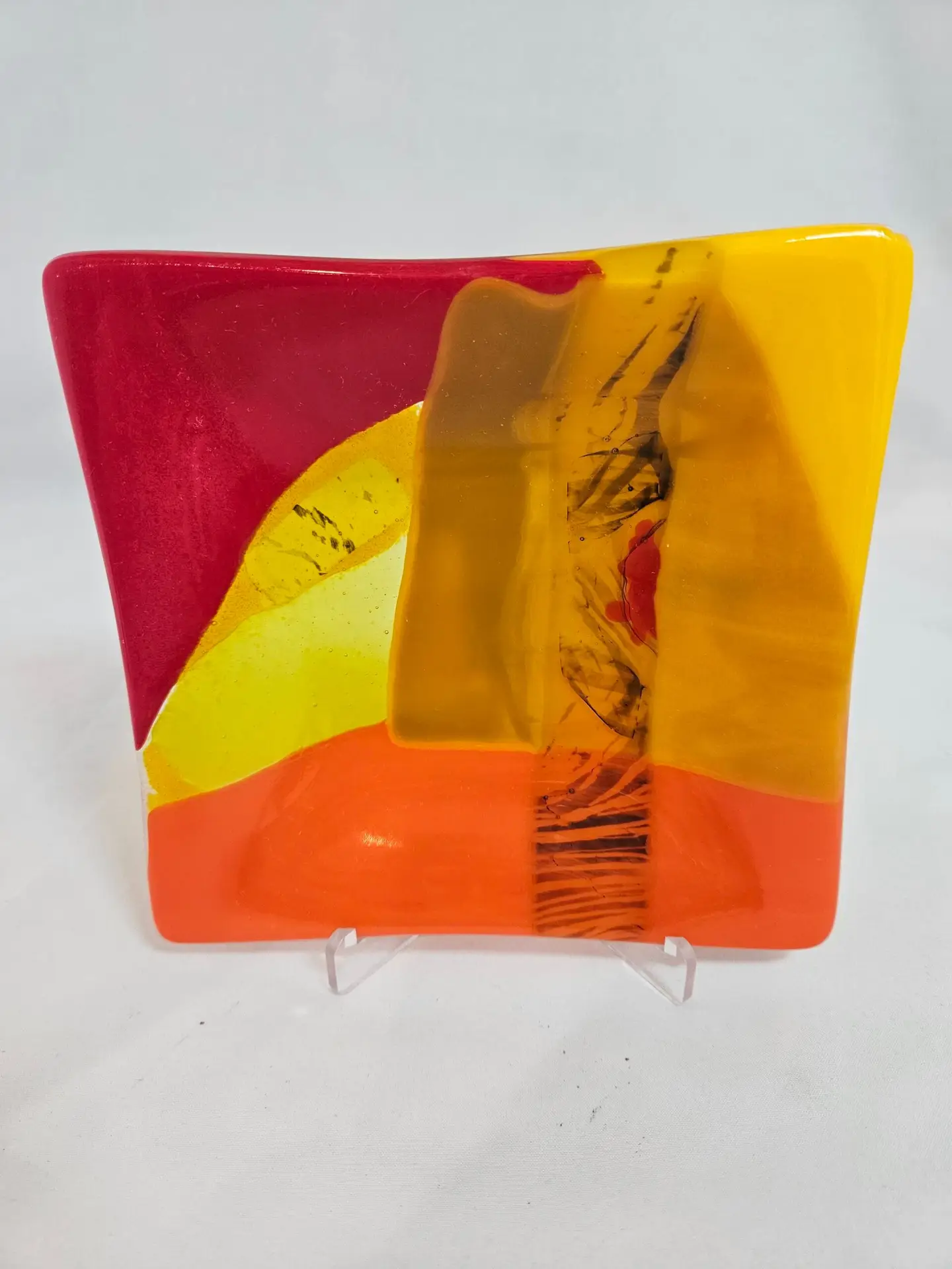 The Radiant Horizon is a vibrant abstract glass sculpture with a wavy shape, featuring red, yellow, and orange sections and a vertical translucent dark streak. Its beautifully displayed on a light stand.