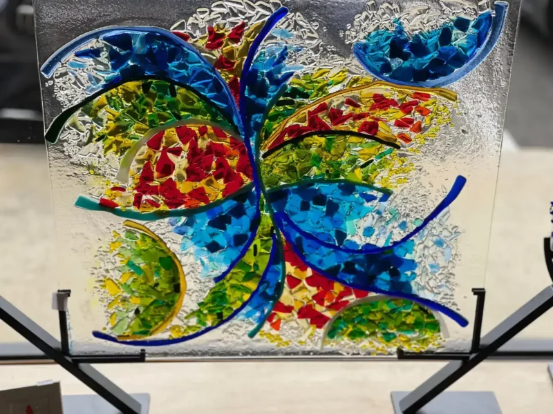Certainly! Here is your text with the background, surface, stand, position, and things near or beside the product removed: The Joy In Motion fused glass sculpture features abstract colorful patterns in blue, red, yellow, and green on a square panel.