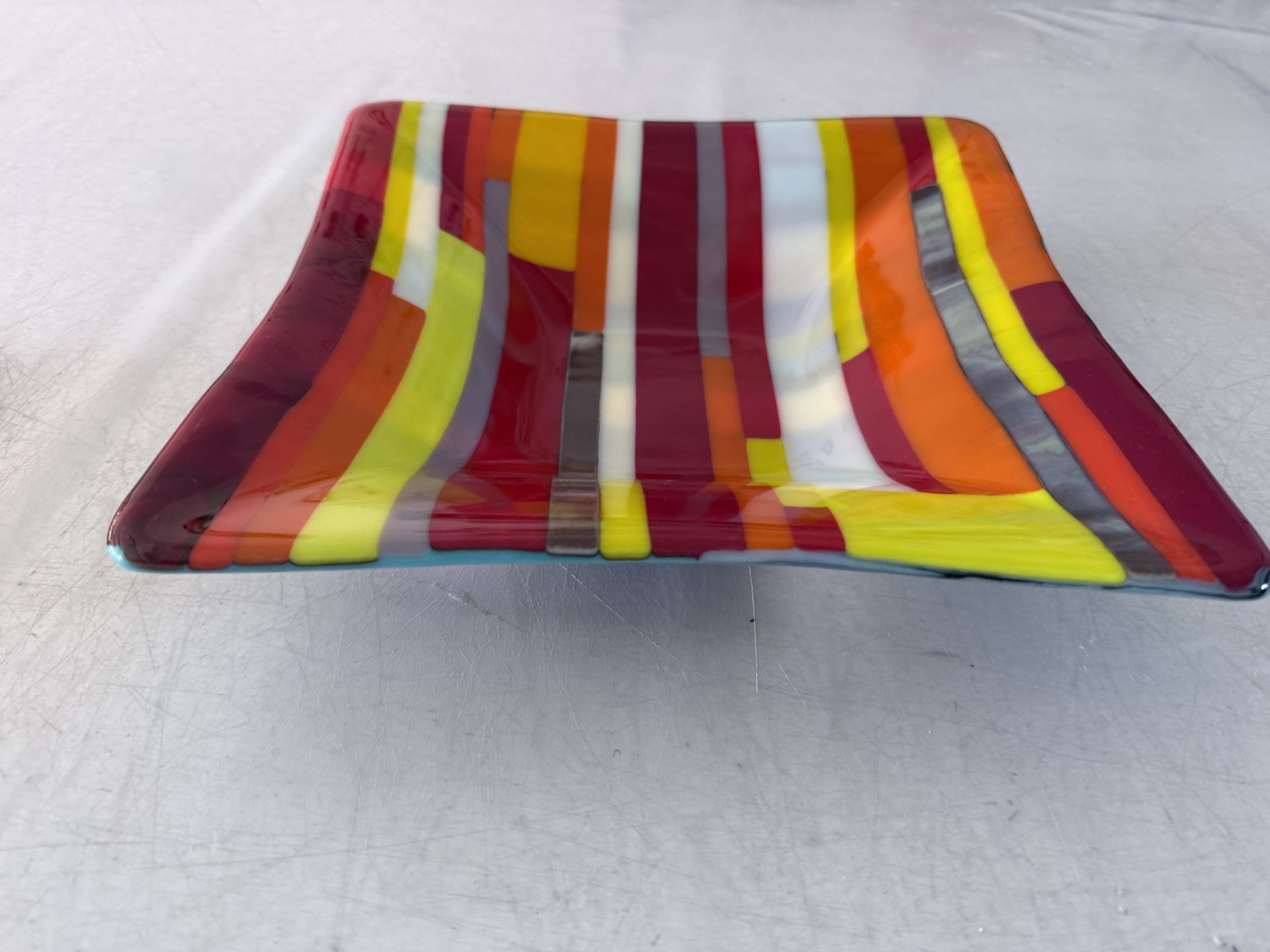 Wider view of the beautiful reds, oranges, yellow and white strips that make up the inside of this square fused glass bowl.