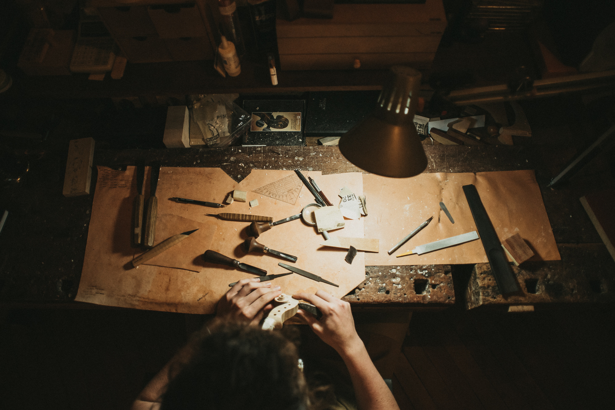 A violin soundpost placed on a luthier's workbench alongside specialized tools like a soundpost setter, chisels, and clamps. The finely carved spruce post rests near the violin's belly, demonstrating precision craftsmanship in instrument adjustment.
