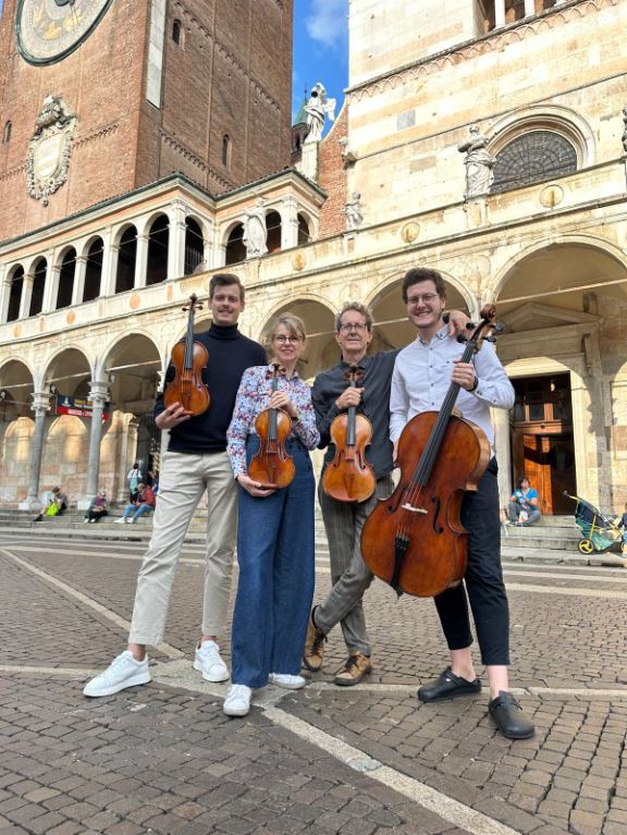 Four luthiers holding string instruments (violins and cello) in front of the historic Duomo of Cremona, Italy — a city renowned for its violin-making tradition.