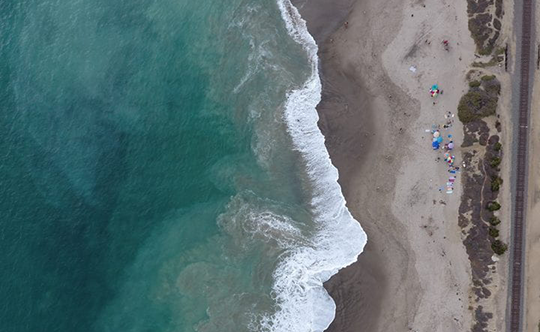 A bird's eye view of San Clemente State Beach in Southern California shows Pacific Ocean waves lapping onto shore, a cluster of umbrellas and, to the far right, a rail corridor that serves passenger and freight trains. UC Irvine's Brett Sanders says San Clemente is one of many examples of beaches in California that demonstrate their value in multiple ways. UC Irvine Flood Lab