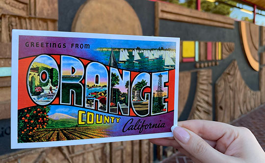 A photo taken of Mexican artist J. Sergio O’Cadiz Moctezuma mural at Fremont Elementary School with an Orange County postcard in front of it. 