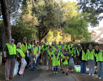 A group of volunteers in yellow vests stand at a cleanup site. A few are holding orange trash grabbers and trash bags.