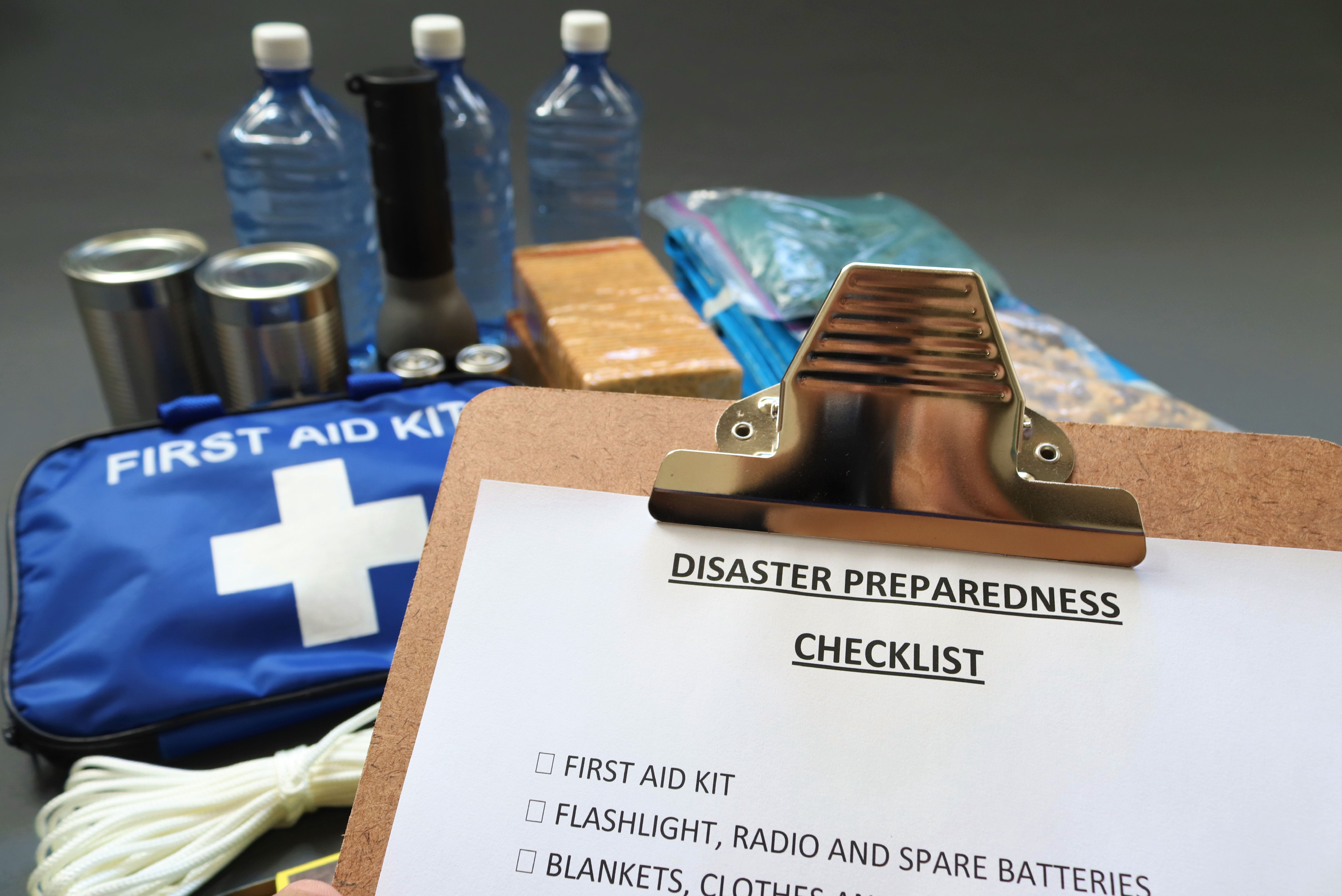 A photo of emergency items sitting together on a surface, including bottled water, a flashlight, canned food, rope, a bag labeled "first aid kit", and other items. In the foreground is a clipboard with a paper that reads "Disaster Preparedness Checklist". Visible items on the checklist are "first aid kit", "flashlight, radio and spare batteries" and "blankets, clothes"