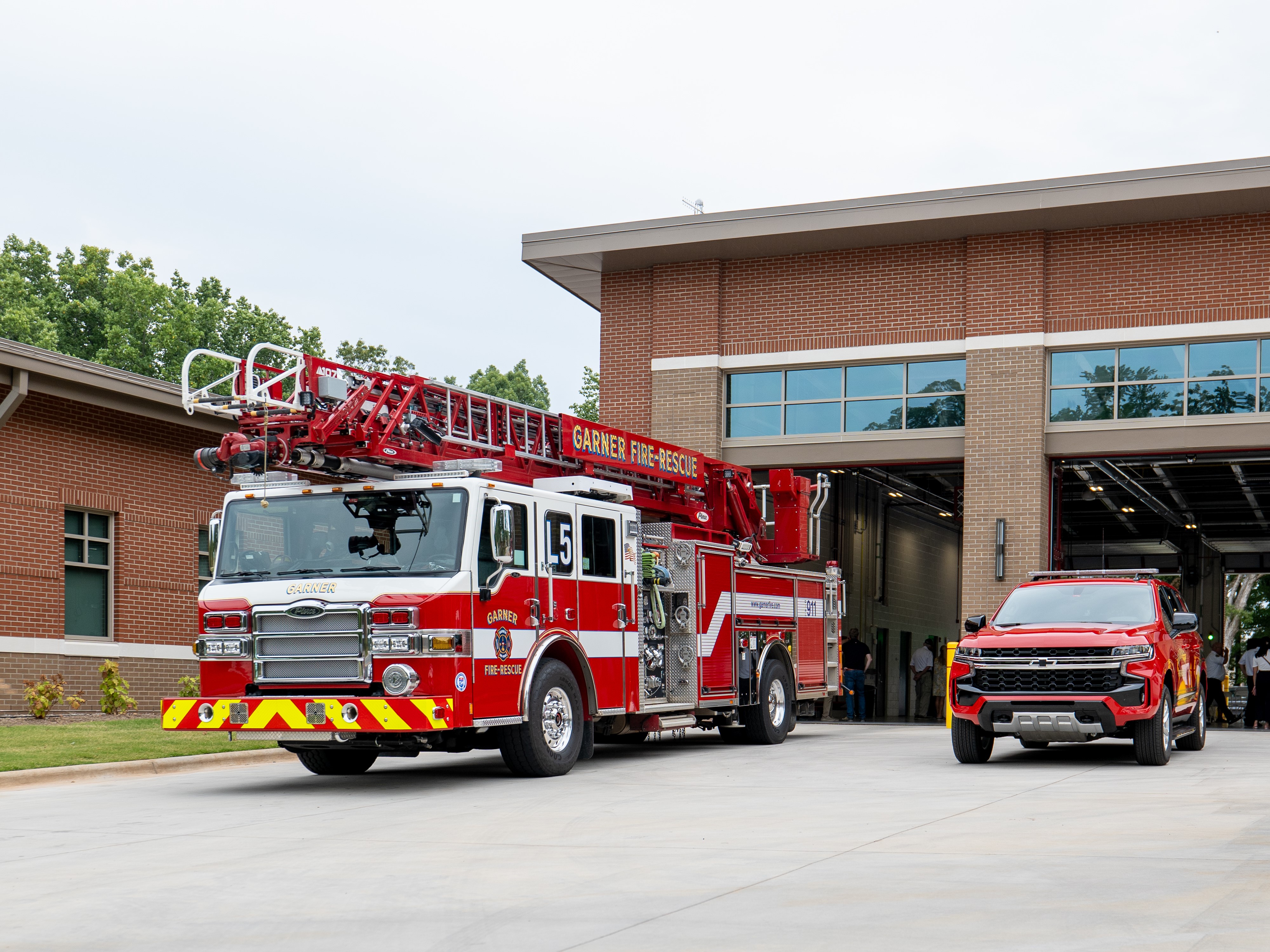 A red Garner Fire-Rescue Department Ladder Truck sits in front of the open bays of a fire station, next to a red SUV.