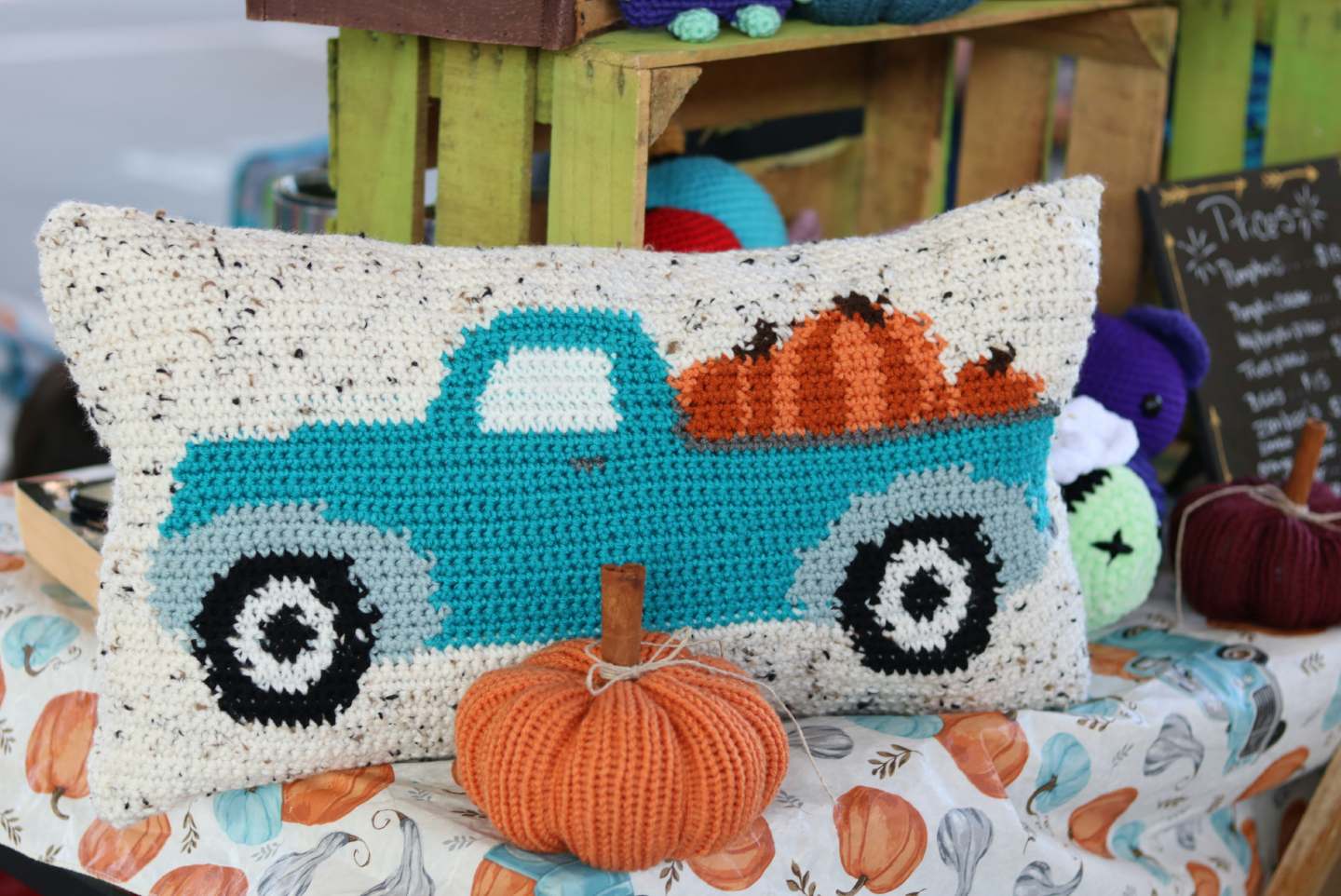 Table with crates and fall table cloth holding a crochet pillow of a teal truck loaded with pumpkins sitting behind a a knitted pumpkin.