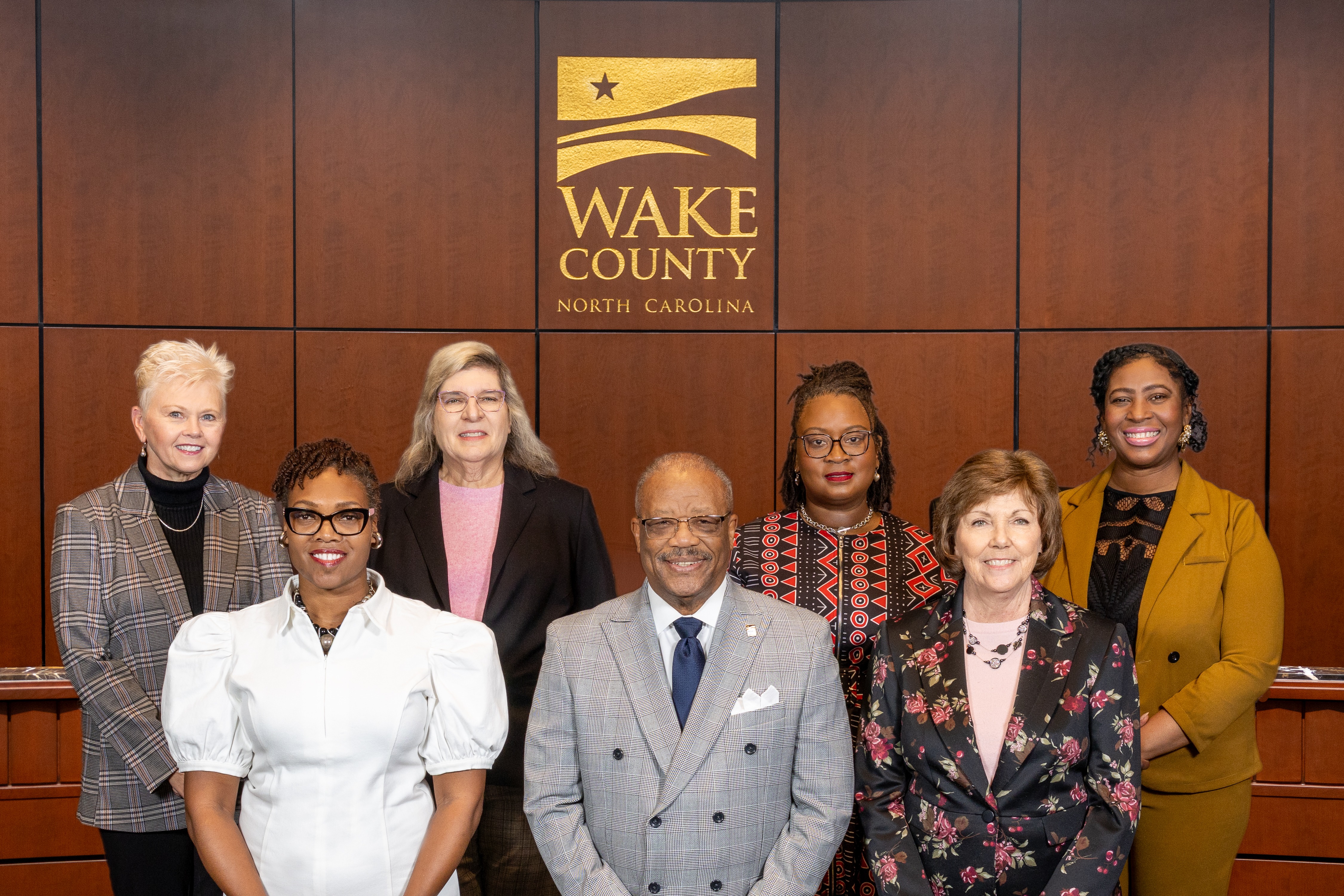 members of the 2026 Wake County Board of Commissioners pose in the Board room under a Wake County logo