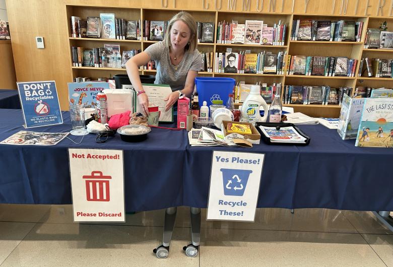 A woman sets up a table with examples of recyclable and nonrecyclable items. Signs on the table read “Not Accepted! Please Discard” on the left and “Yes Please! Recycle These!” on the right. The display includes bottles, cardboard and food containers.