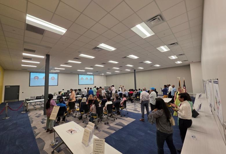 Group of people walking and sitting in a room with tables and signs. Images are projected on the walls.