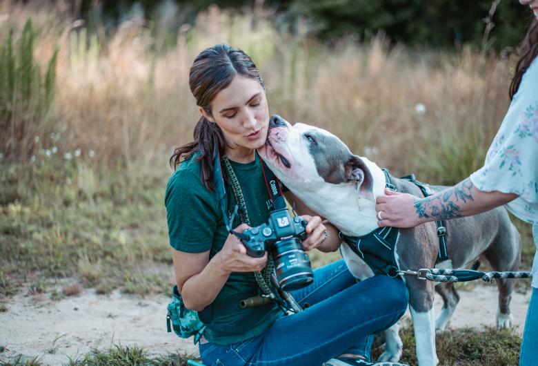 A woman sitting outdoors holds a camera while a large gray-and-white dog affectionately licks her face. Another person with a floral tattoo on their arm holds the dog’s leash. The scene takes place in a grassy field with warm, natural light.