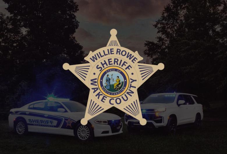 Badge-shaped logo for the Wake County Sheriff’s Office featuring the North Carolina state seal and the name “Willie Rowe, Sheriff, Wake County.” Behind the badge are two sheriff’s vehicles with blue lights on, parked on grass with trees and a dusk sky in the background.