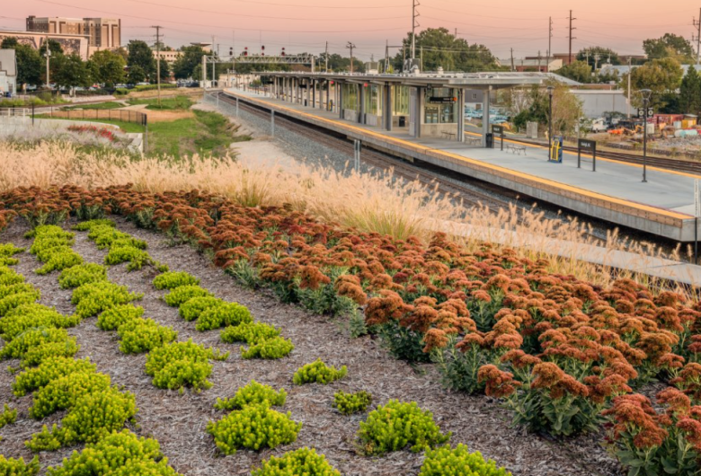 A landscaped area with colorful, low-growing plants and tall grasses in the foreground overlooks a modern train platform with glass shelters and yellow-edged platforms. The scene is set at sunset with warm light, and city buildings and trees are visible in the background.