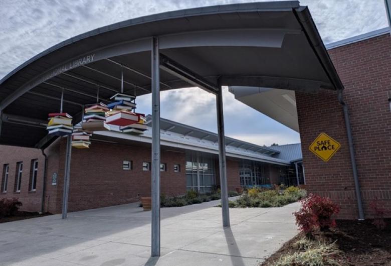 Entrance of a public library with a covered walkway and a hanging sculpture of colorful books. A yellow ‘Safe Place’ sign is mounted on the brick wall.