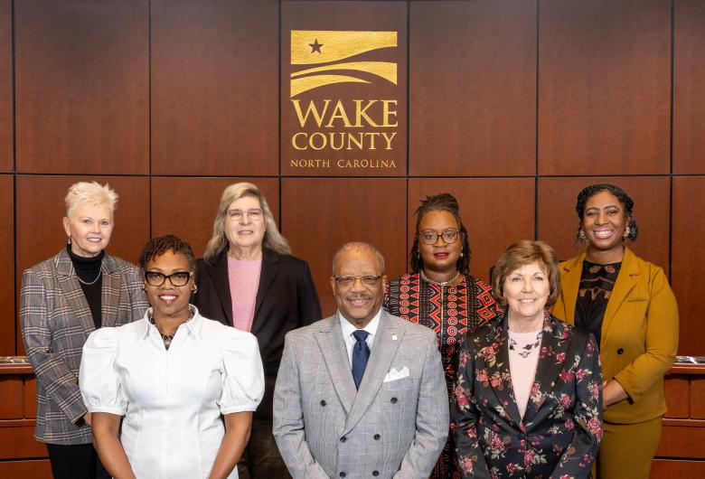 members of the 2026 Wake County Board of Commissioners pose in the Board room under a Wake County logo