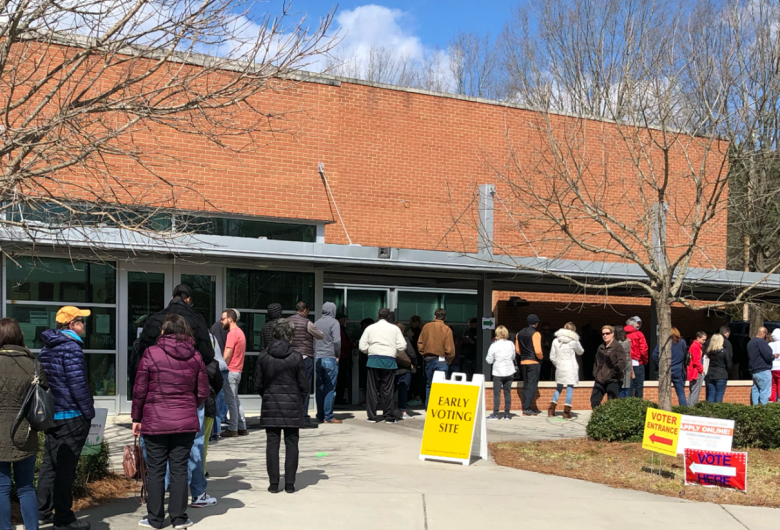 **Alt text:** People standing in line outside a brick building marked as an early voting site.