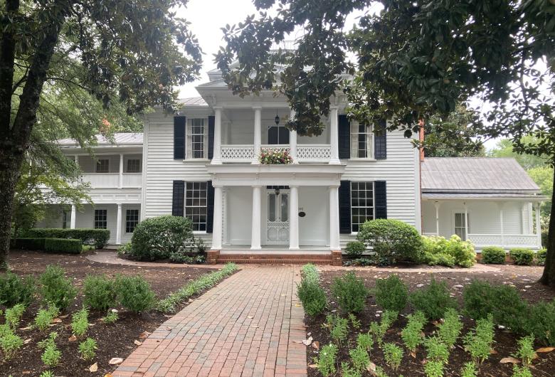 Large white historic home with black shutters, tall columns, and a brick walkway leading to the front entrance, surrounded by trees and landscaped gardens.