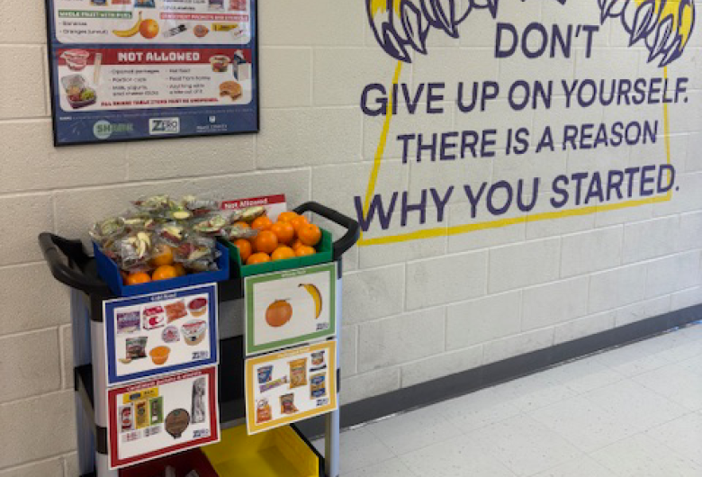 Project SHARE food cart with fruit and packaged snacks beneath a “Share” food donation poster, next to a wall mural of a tiger and the message “Don’t give up on yourself. There is a reason why you started.”