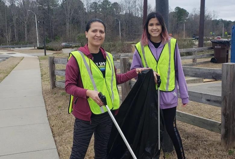 Two volunteers wearing safety vests collect roadside litter with grabbers and a trash bag along a sidewalk during a community cleanup.