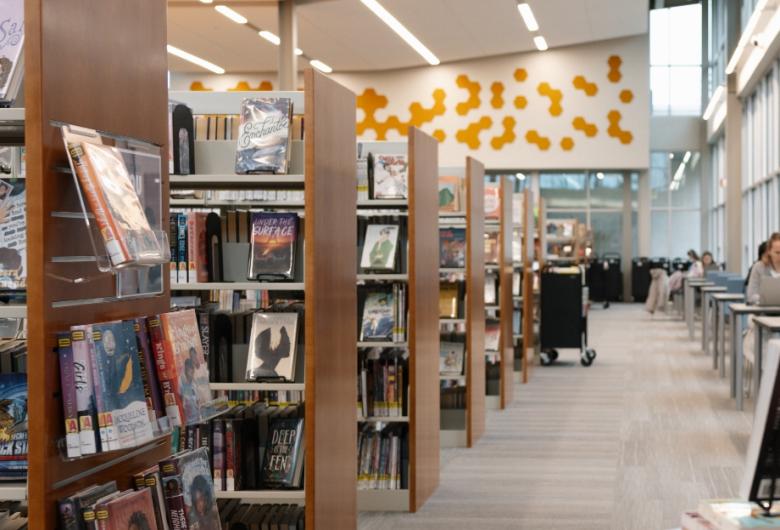 Rows of bookshelves inside a modern library, with patrons reading and working at tables along large windows.
