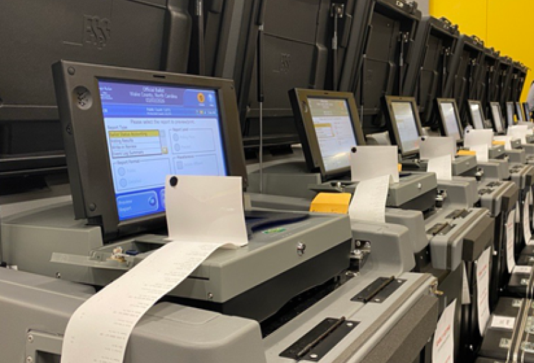 Row of voting machines with screens and printed ballots in a polling or election processing area.