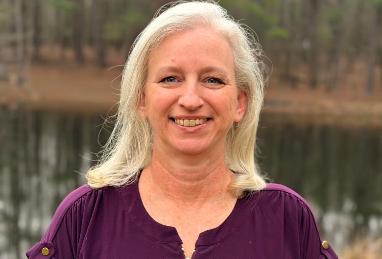 A smiling woman with light hair wearing a purple top, standing outdoors near a calm body of water with trees in the background.