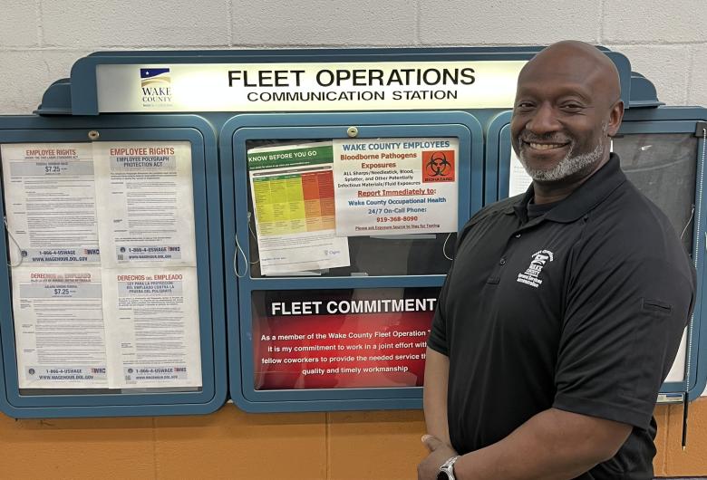 Man stands smiling beside a “Fleet Operations Communication Station” board displaying employee notices.