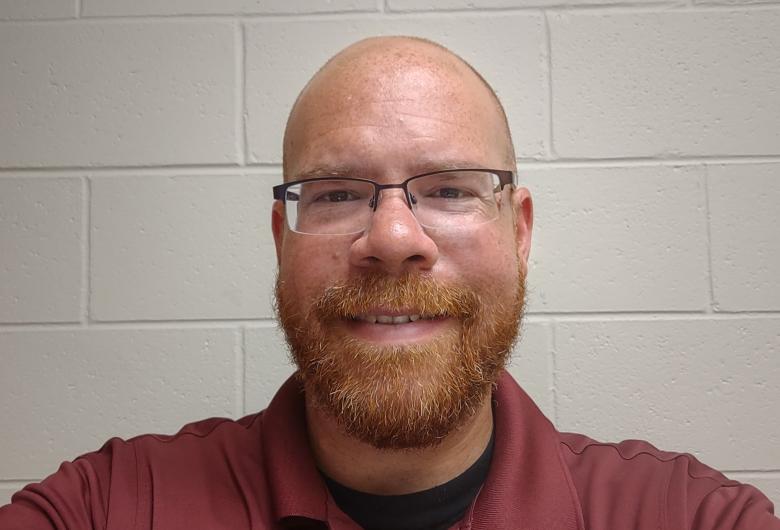 Man with glasses and a beard smiles at the camera against a plain wall, wearing a Wake County General Services shirt.