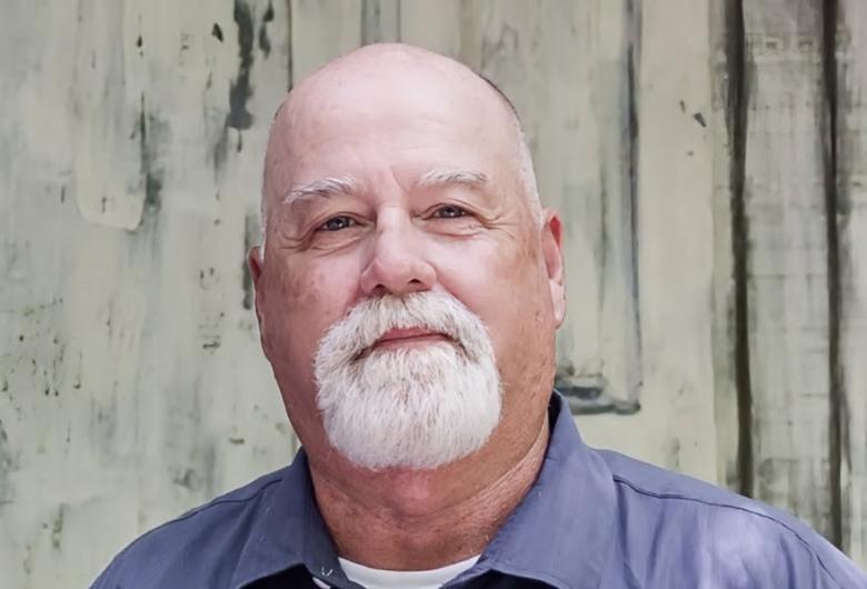 Older man with a bald head and white beard, wearing a blue button-up shirt, standing in front of a weathered wall.