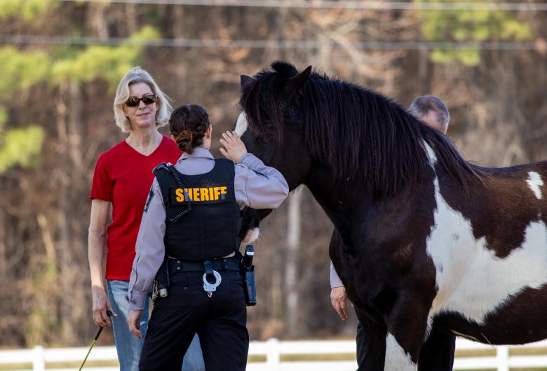 WCSO deputy petting a brown and white horse. A woman and man are standing next to the horse, facing the camera. Woods and a white picket fence are in the background.