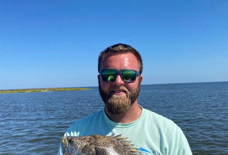 Man on a boat holding a large fish, with water and shoreline in the background.