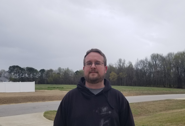 Man wearing glasses and a hoodie stands on a driveway in a residential area under a cloudy sky.