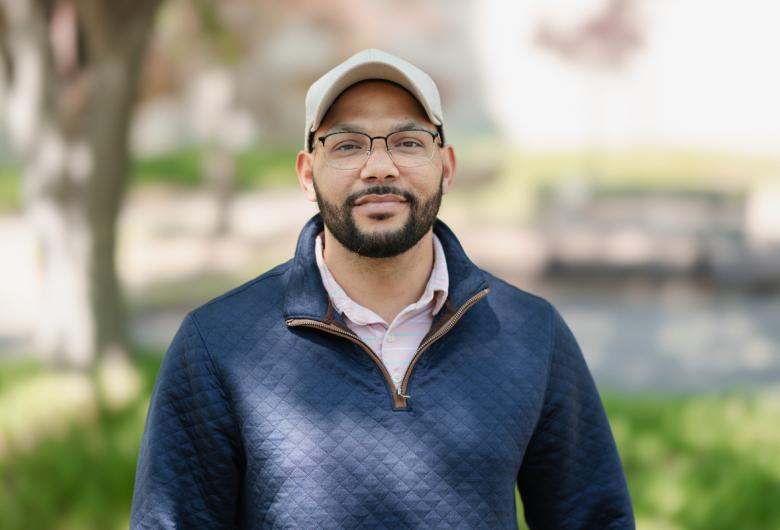 Smiling man with a cap, glasses and beard, wearing a thermal pullover, stands in a landscaped parking lot.