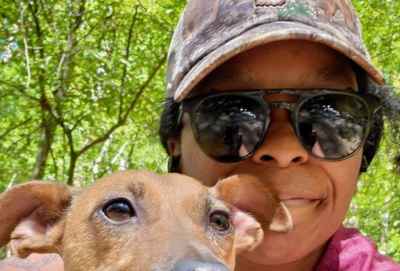 Person wearing sunglasses and a cap poses with a small dog in a blue harness on a wooded trail, with green trees in the background.