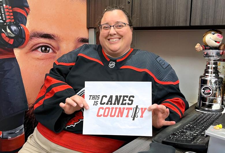 Smiling person in a Carolina Hurricanes jersey holds a “This Is Canes Country” sign while seated at an office desk with Hurricanes-themed decor.