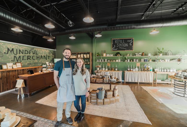 **Alt text:** Two people stand smiling inside a refill and sustainable goods store, surrounded by shelves of reusable containers, household products, and eco-friendly merchandise.