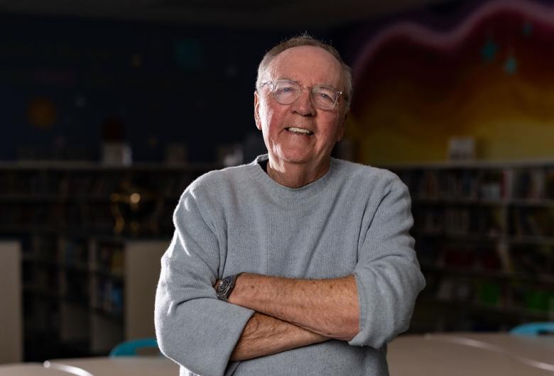 Author James Patterson, wearing light gray sweater, stands with arms crossed in a library.
