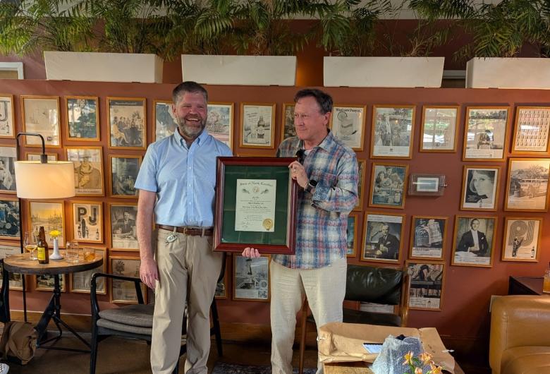 Two men stand together while one presents the other with a framed Order of the Long Leaf Pine certificate during an indoor recognition event.