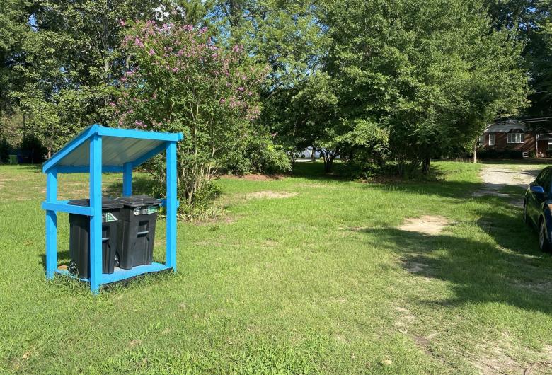 Bright blue shelter holding two trash carts beside a grassy driveway surrounded by trees.