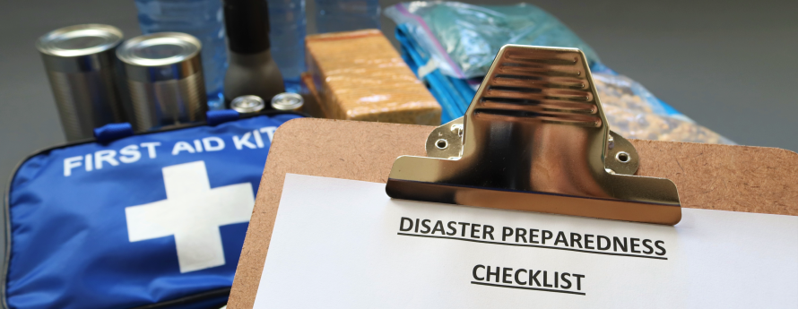 A photo of emergency items sitting together on a surface, including bottled water, a flashlight, canned food, rope, a bag labeled "first aid kit", and other items. In the foreground is a clipboard with a paper that reads "Disaster Preparedness Checklist". Visible items on the checklist are "first aid kit", "flashlight, radio and spare batteries" and "blankets, clothes"