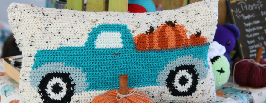 Table with crates and fall table cloth holding a crochet pillow of a teal truck loaded with pumpkins sitting behind a a knitted pumpkin.