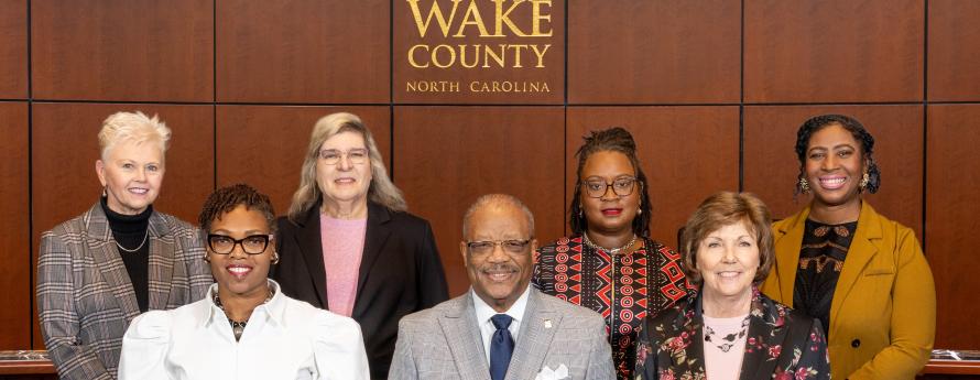 members of the 2026 Wake County Board of Commissioners pose in the Board room under a Wake County logo