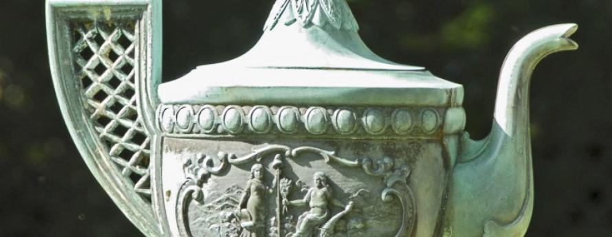 Bronze teapot-shaped drinking fountain on a pole in a park with a wrought-iron fence and distant water view.