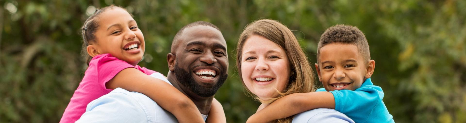 Two adults smile while holding children on their shoulders outdoors.