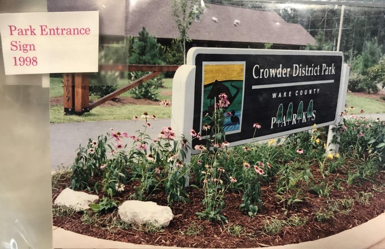 old picture from 1998 of first crowder park front entrance sign surrounded by flowers with park office in background