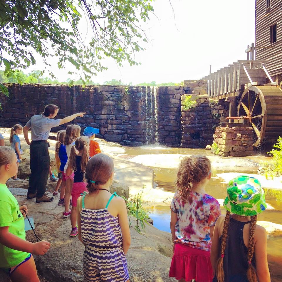 Group of children stand in front of mill dam and waterfall with mill building off to the side