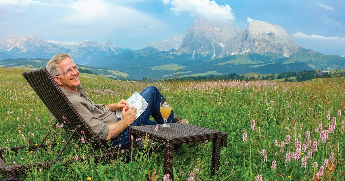 TV host Rick Steves smiles from a wicker lounge chair in a field, a map in his lap, mountains in the background